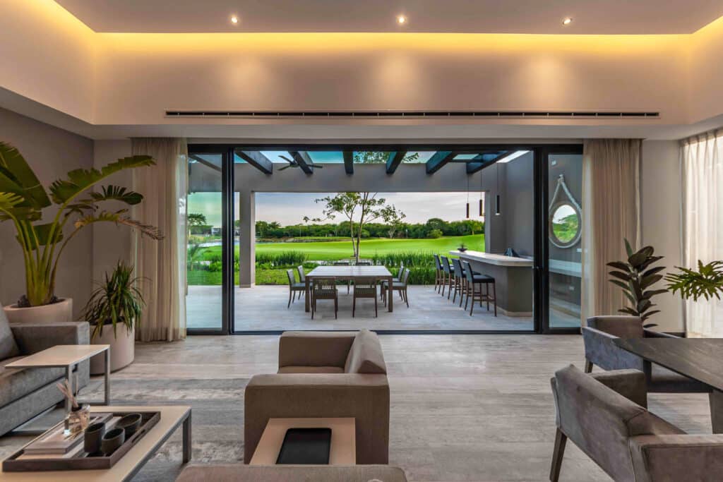 A spacious living room with large sliding glass doors opening to a covered patio overlooking green grass and trees in Yucatan Country Club, Merida.