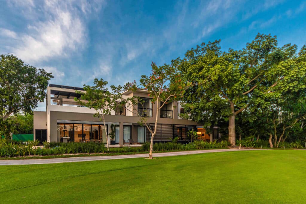 A modern two-story house with large glass windows, surrounded by lush greenery and trees at Amanha A in Yucatan Country Club, Merida.