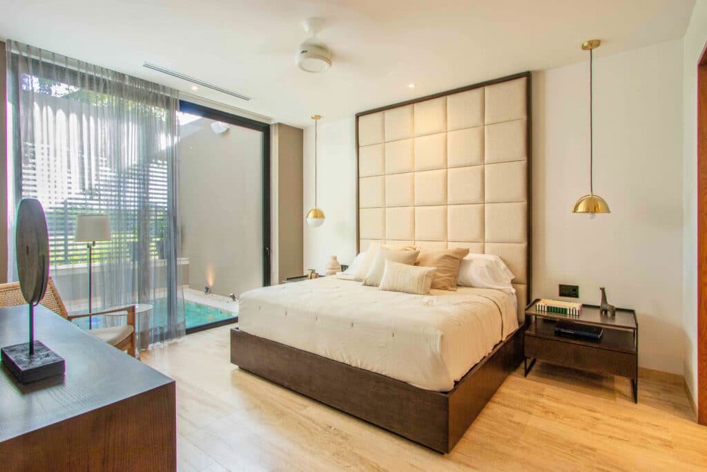 Modern bedroom in the Amanha A-16 development at Yucatan Country Club, Merida, with a large bed, beige upholstered wall, and sliding glass door to a small outdoor space.