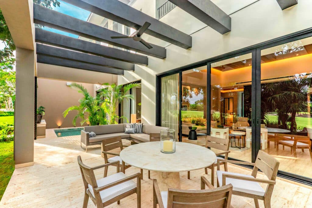 A modern outdoor living space at Amanha A-16 in Merida's Yucatan Country Club features a covered patio with a round dining table, a seating area, lush greenery, and large glass doors opening to the interior.