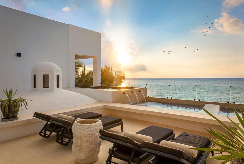 Beachfront view at Bahia Tankah 30 in Tulum shows a terrace with lounge chairs, a small pool, and the ocean with a sunrise sky and some birds flying overhead.