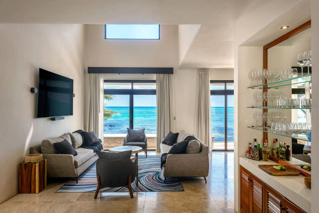 Interior view of Bahia Tankah 30 in Tulum, featuring a beachfront living room with large windows overlooking the ocean, a TV, and a bar area with glassware.