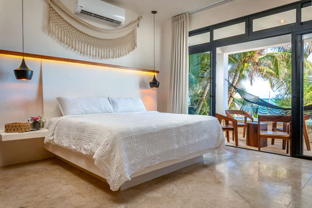 A bedroom at Bahia Tankah 30 in Tulum features a white bed, a balcony with a hammock, wooden chairs, tropical trees, and a view of the beach.