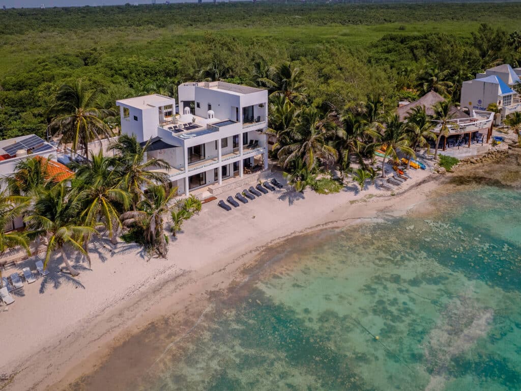 An aerial view of Bahia Tankah 30 beachfront property in Tulum, featuring a white modern house surrounded by palm trees, with a sandy shoreline and clear turquoise waters.