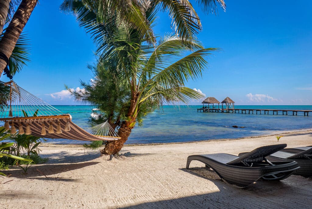 Bahia Tankah 30 in Tulum features a beachfront setting with a sandy shore, palm trees, a hammock, lounge chairs, and a pier extending into the clear blue water.