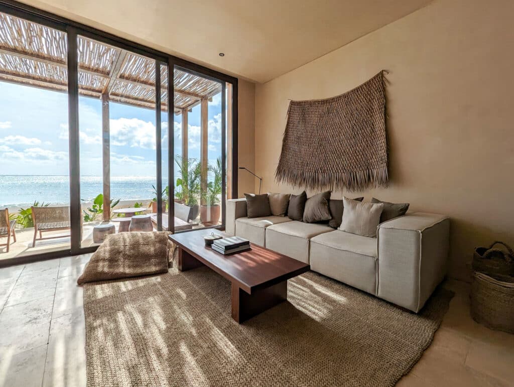 Living room with a beige sofa and dark pillows facing large glass sliding doors that open to a beachfront view with sand, greenery, and ocean, in Bahia B development, Tulum.