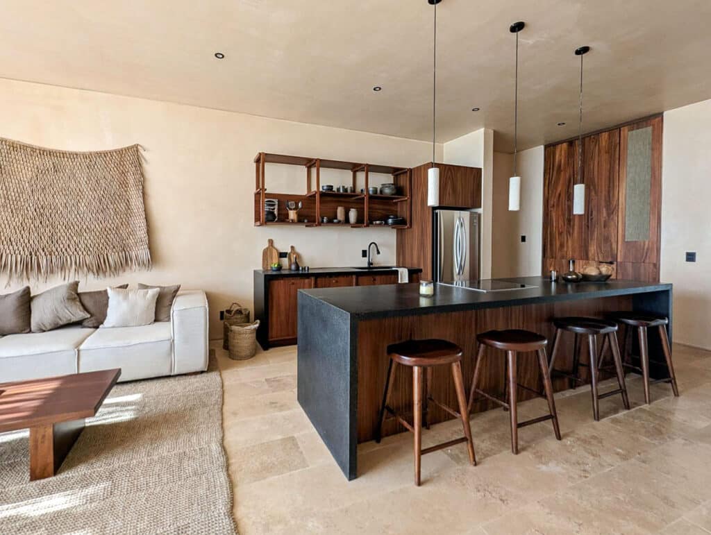 Interior view of Bahia B-31 in Tulum, featuring a modern kitchen with wooden accents, open shelving, a black countertop island, and adjacent beige living area.