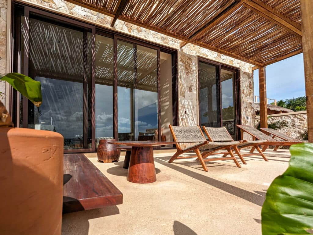 A spacious beachfront terrace at Bahia B in Tulum features wooden lounge chairs, a small table, and a natural stone wall with large sliding glass doors and a bamboo ceiling.