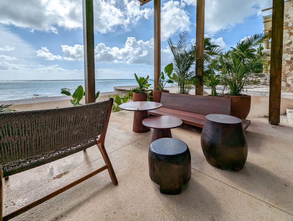 View of a beachfront terrace at Bahia B in Tulum featuring wooden seating, tables, lush potted plants, and a scenic ocean backdrop with clouds in the sky.