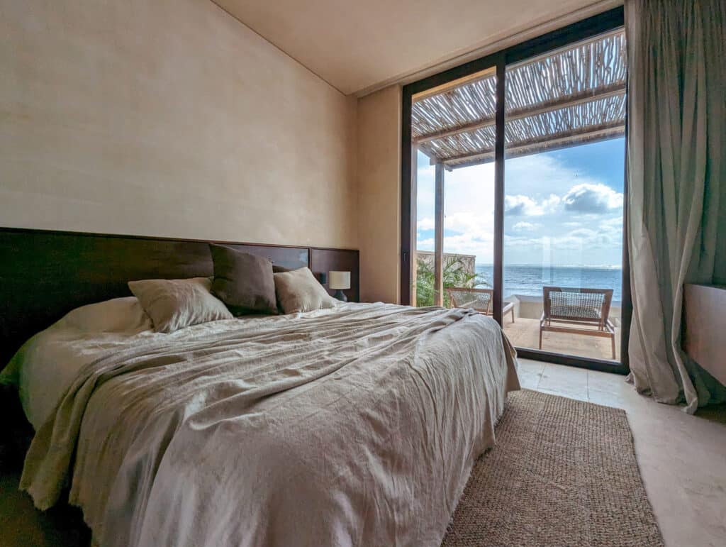 A bedroom in Bahia B, Tulum, with a large sliding glass door opening to a balcony overlooking the beach and ocean, featuring neutral-toned bedding and a woven rug.
