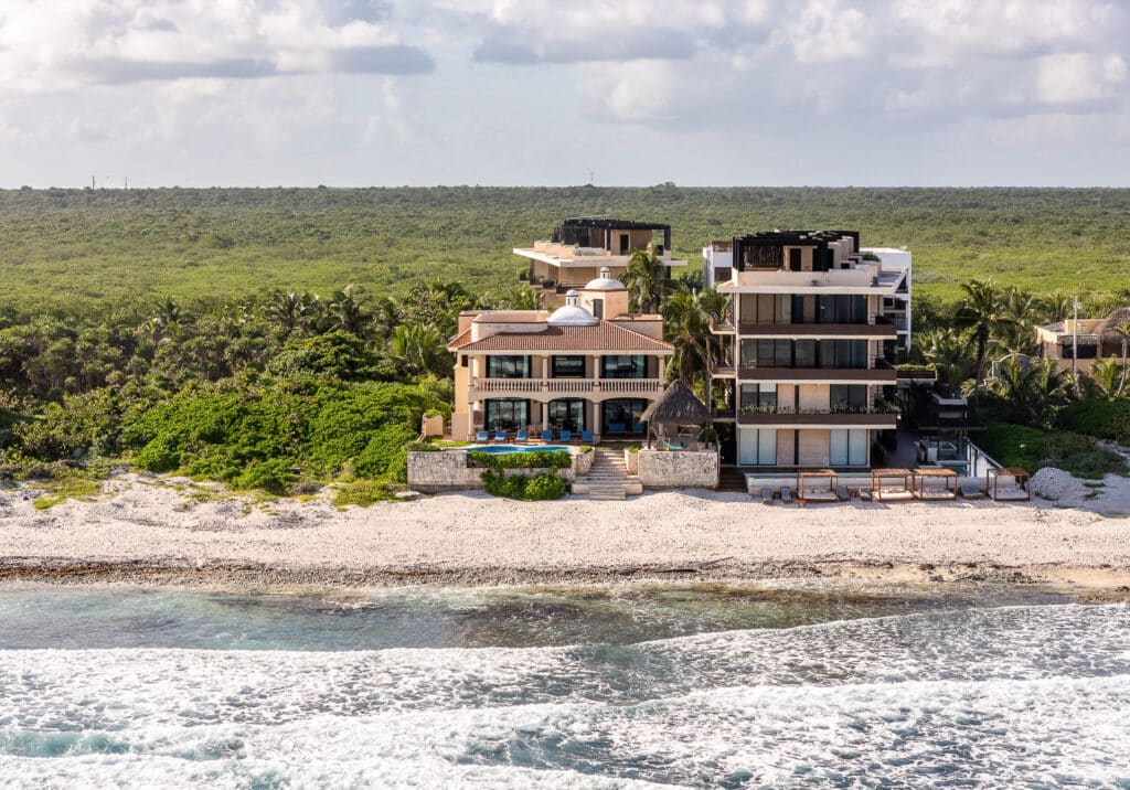 A modern beachfront residence at Casa Tres Palmeras in Tankah Bay, Tulum, with multiple terraces, lush greenery, and views of the shoreline under a cloudy sky.
