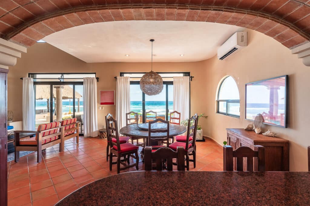 Interior view of Casa Tres Palmeras in Tankah Bay, Tulum, showing a dining area with a round table, chairs, and large windows with ocean views.
