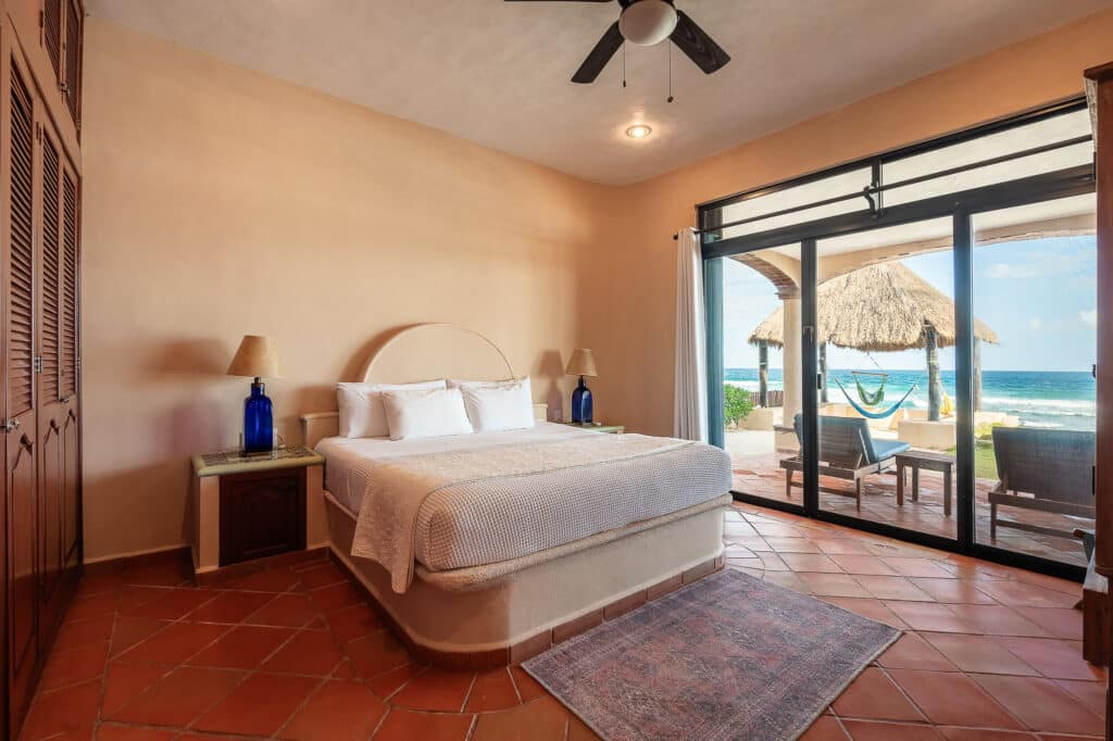 Bedroom interior with a large sliding door opening to a beachside patio featuring lounge chairs, a thatched gazebo, and a hammock in Tulum.