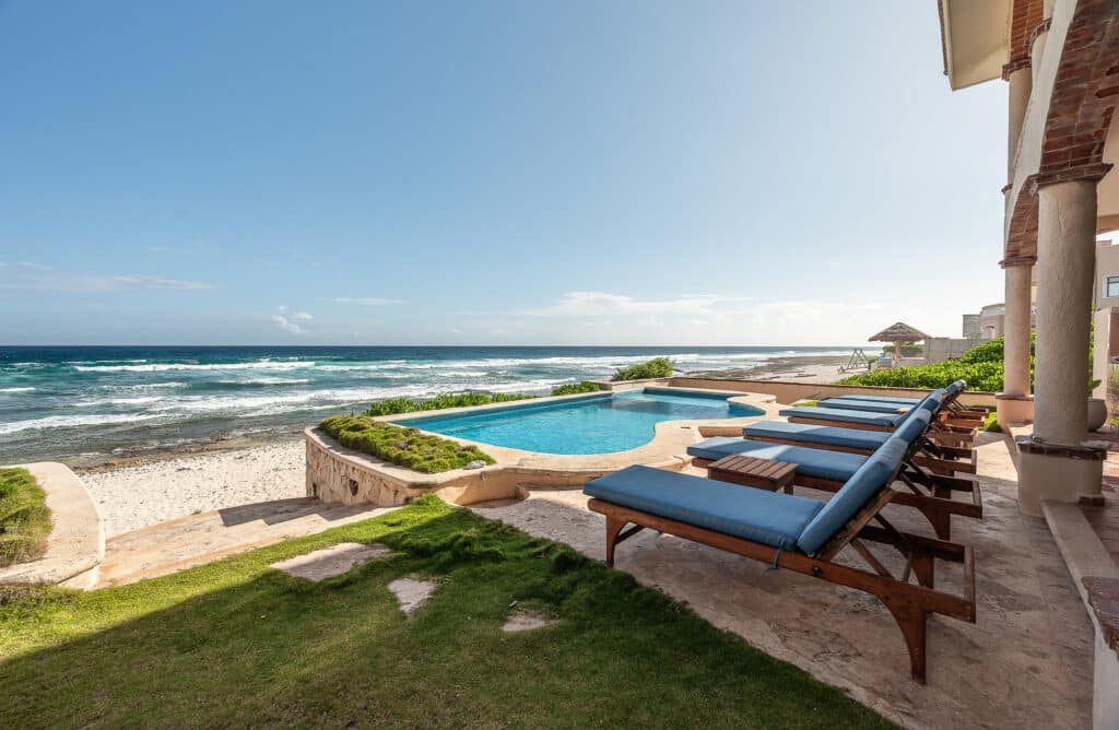 A modern house with an inviting pool overlooks the sandy beach and ocean, featuring lounge chairs and lush greenery at Casa Tres Palmeras in Tankah Bay, Tulum.