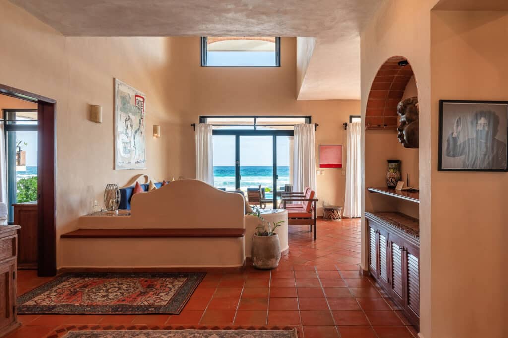 Living room in Casa Tres Palmeras, Tankah Bay, Tulum, featuring ocean views through large glass doors with white curtains. The room has terracotta tile flooring and warm-colored walls.