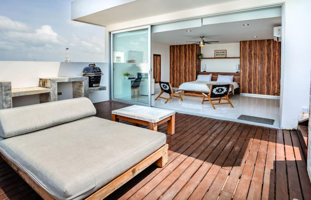 A penthouse unit at DK05 – 401 in Playa del Carmen features a spacious outdoor terrace with a lounge area, and an interior bedroom with modern decor and wood accents.