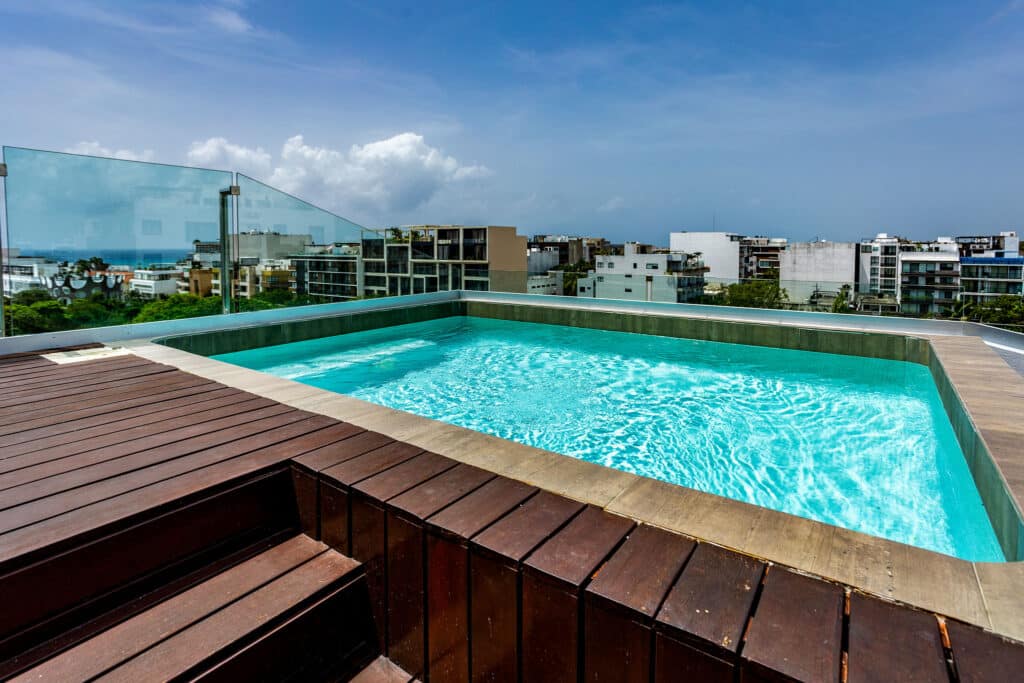 A rooftop infinity pool offers a view of Playa del Carmen skyline in the DK05 – 401 development in Luis Donaldo Colosio, with glass railings and cityscape background.
