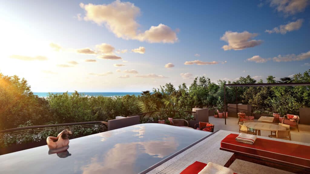 A spacious balcony at Faena District B-2303 in Tulum, offers lush greenery and views of the ocean, with an infinity pool, potted plants, and seating areas in a tropical setting.