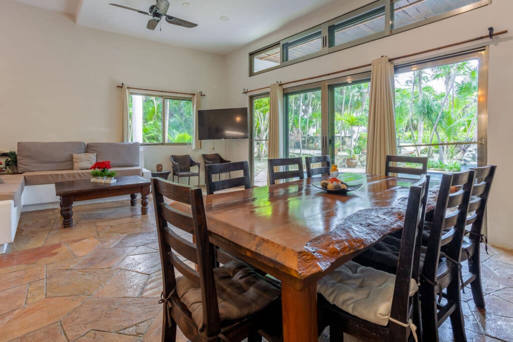 Living room in Autumns Retreats at Tankah Bay, Tulum, featuring large windows with views of lush greenery, a wooden dining table, and a neutral-colored sofa.