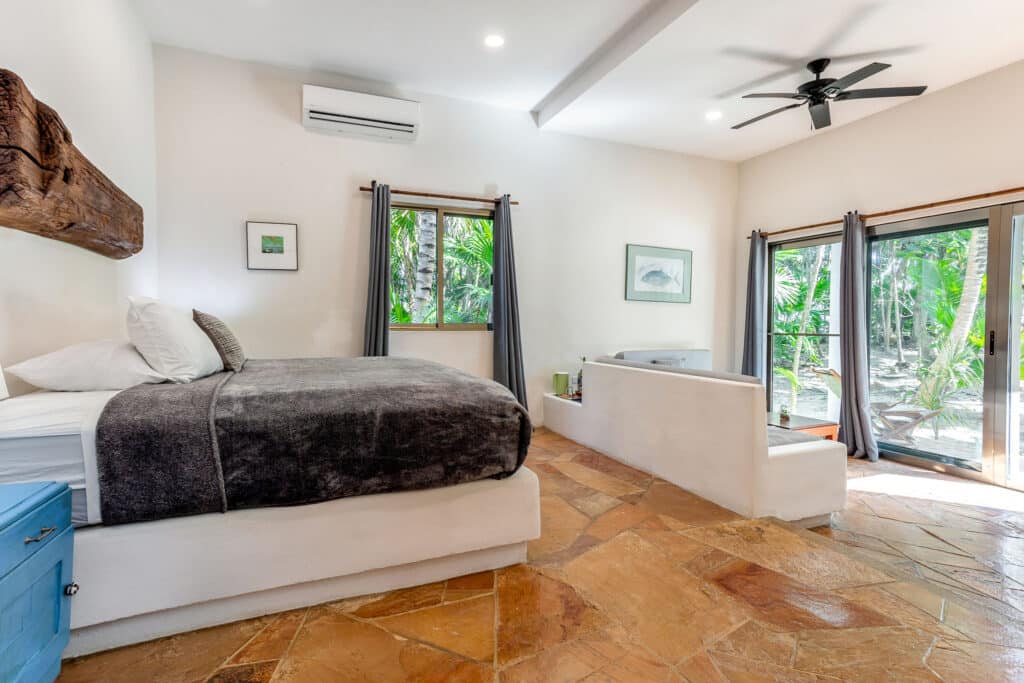Interior view of Villa Eli 4 at Autumns Retreats in Tankah Bay, Tulum, showcasing a bedroom with a stone floor, a bed with a dark blanket, and large windows with views of lush greenery.