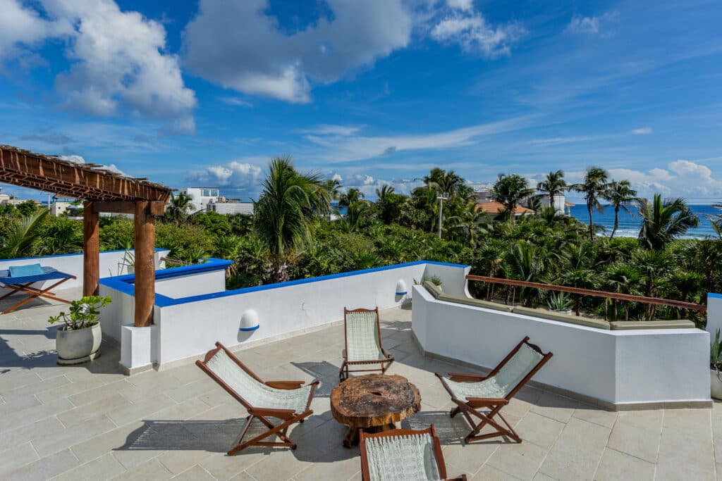 A spacious terrace at Autumns Retreats in Tankah Bay, Tulum, offers seating with a view of lush greenery and the ocean in the distance under a partly cloudy sky.