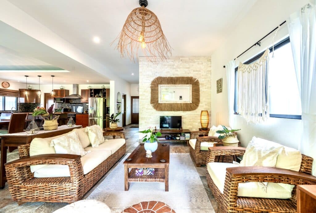 Living room interior in Villa Victoria, Puerto Aventuras, featuring wicker furniture, a white brick fireplace, natural light from windows, and tropical decor.