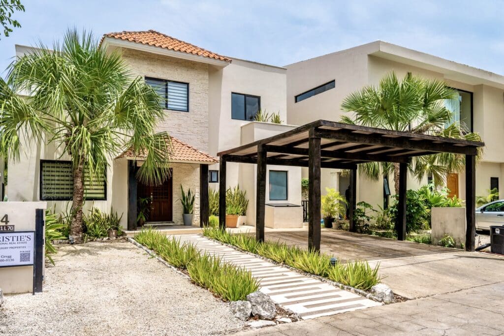 Exterior view of Villa Victoria in Puerto Aventuras, featuring a modern design with a covered parking area, lush tropical plants, and a landscaped entrance along the golf course.