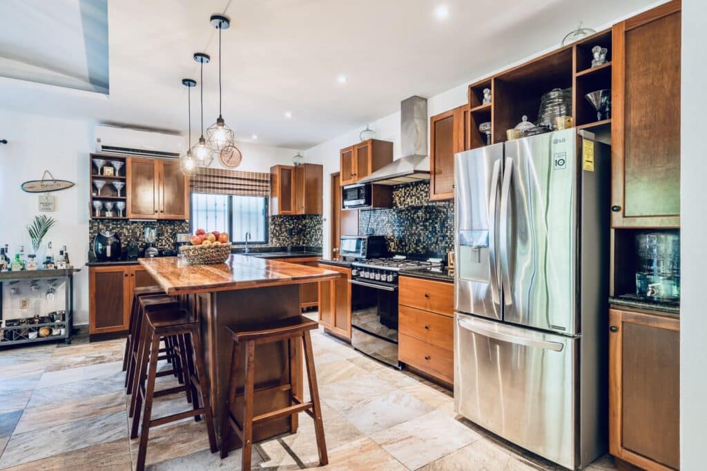 Interior view of a kitchen in Villa Victoria, Puerto Aventuras, featuring wooden cabinetry, a stainless steel refrigerator, a central wooden island, and mosaic tile backsplash.