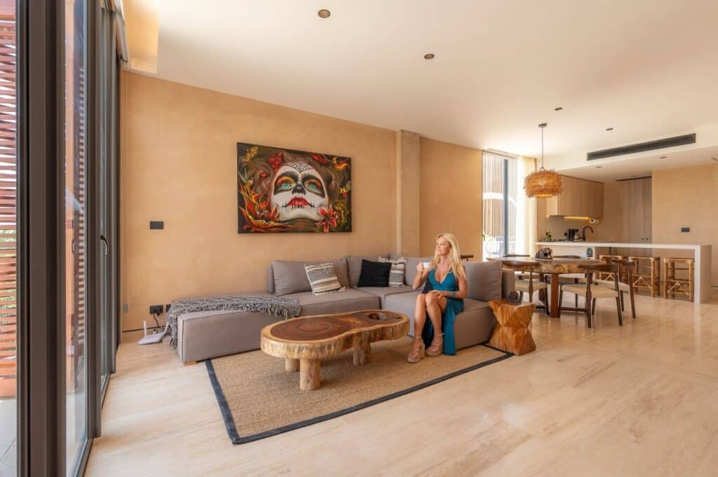 A woman sits on a gray sofa in the living area of The Village 306 penthouse in Corasol, Playa del Carmen, with a wooden coffee table and modern decor, including colorful artwork on the wall.