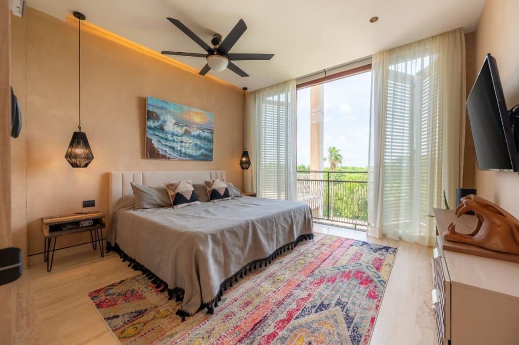 A modern bedroom in The Village 306 penthouse at Corasol in Playa del Carmen features a large bed with decorative pillows, a ceiling fan, a colorful rug, and a balcony with lush greenery outside.