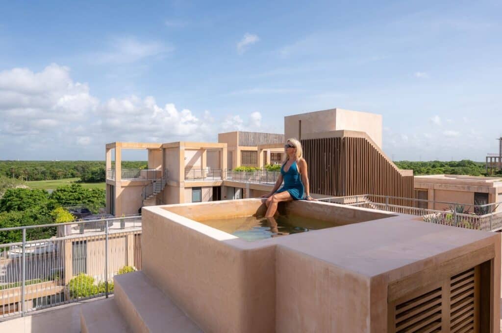 A woman relaxing on a rooftop hot tub in The Village 306 penthouse, located in the Corasol area of Playa del Carmen, with lush greenery and modern architecture in the background.