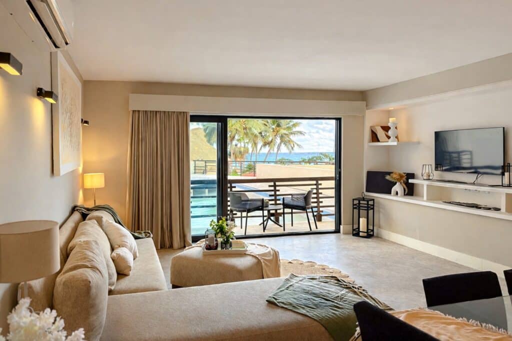 Living room with beige sofa overlooking balcony with outdoor seating and views of palm trees and ocean at Aldea Thai in Playa del Carmen.