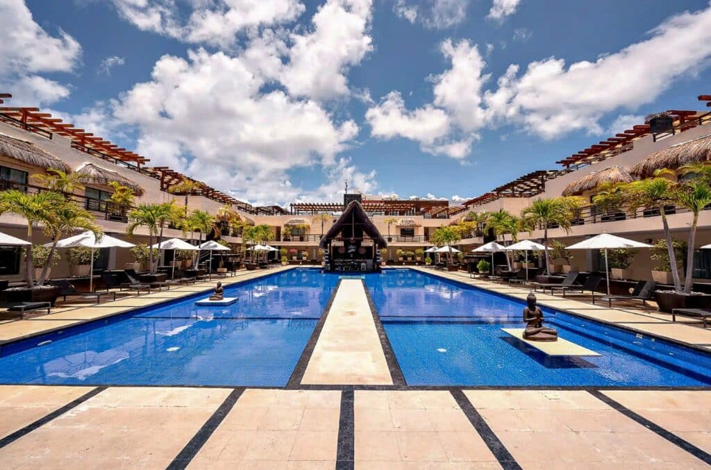 View of a large outdoor swimming pool surrounded by lounge chairs, umbrellas, and tropical trees in a resort courtyard during daytime at Aldea Thai in Playa del Carmen.