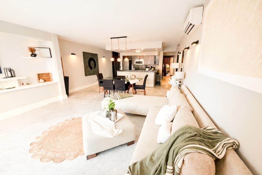 Living room with beige sofa, white walls, and open kitchen with gray cabinetry at Aldea Thai in Playa del Carmen.