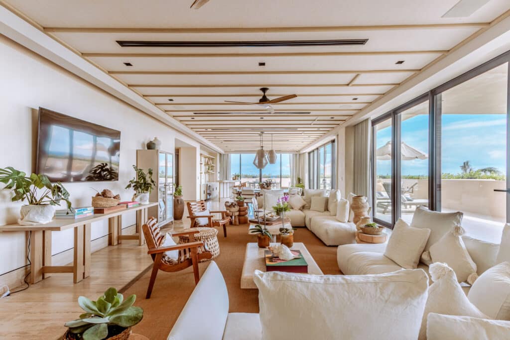 A spacious living room at Costa Residences 1 in Playa del Carmen features white sofas, wooden chairs, large glass doors opening to a balcony with ocean views, and stylish decor.