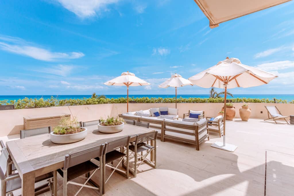 A spacious beachfront terrace at Costa Residences 1 in Playa del Carmen features outdoor seating with umbrellas and overlooks the ocean with clear blue skies.