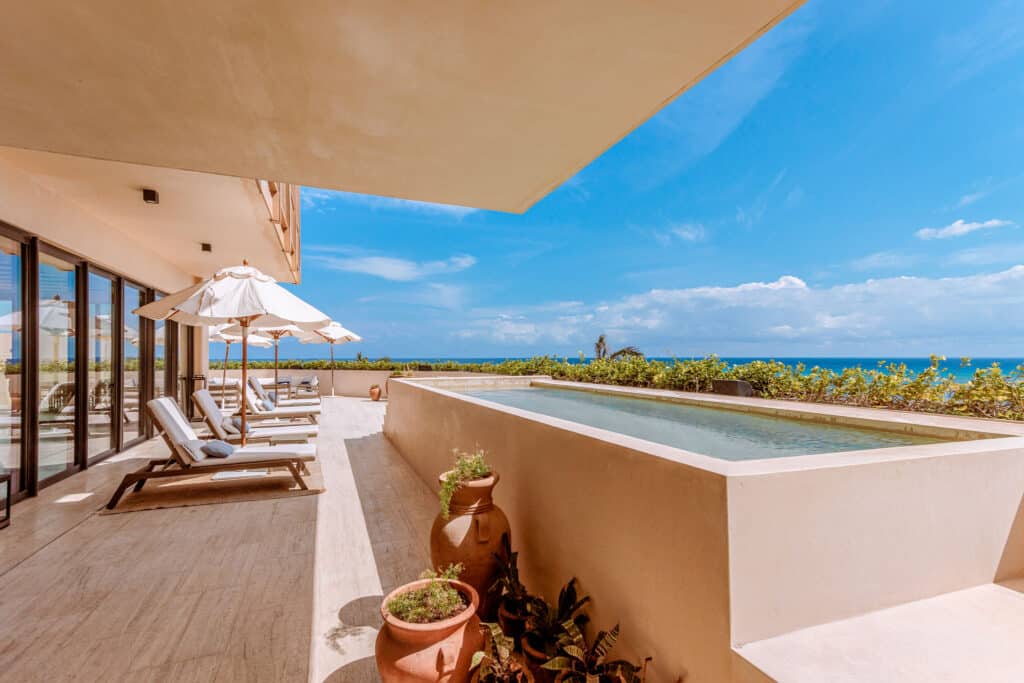A spacious beachfront terrace at Costa Residences 1 in Playa del Carmen features a pool, sun loungers with umbrellas, and a view of the ocean under a clear blue sky.