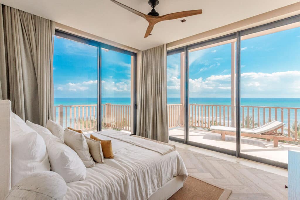 A bedroom with large glass sliding doors opening to a beachfront balcony at Costa Residences 1 in Playa del Carmen, showcasing views of the ocean and sky.