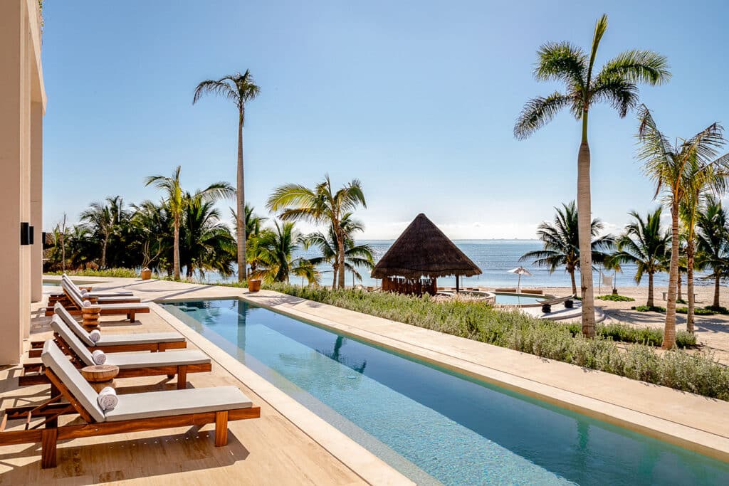 A view from Costa Residences 1-3B in Playa del Carmen shows a beachfront pool area with lounge chairs, tall palm trees, a thatched palapa, and the ocean in the background.