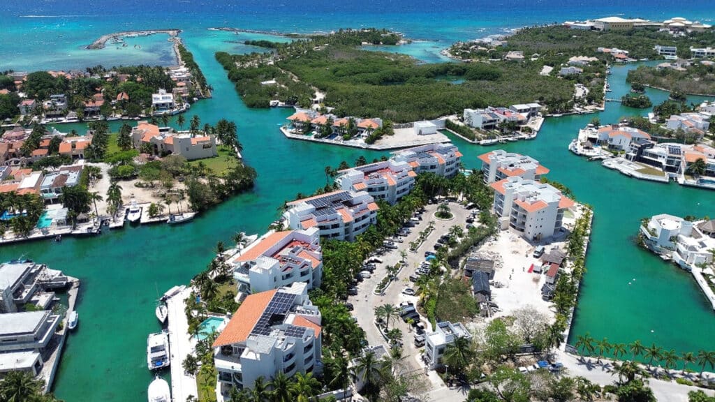 Aerial view of Gran Peninsula I in Puerto Aventuras showing modern apartment buildings along marina front with boats, lush greenery, and clear blue waters surrounding the area.