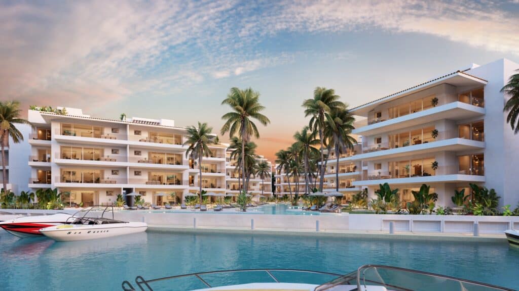 Modern white residential buildings in Marina Aqua A on Puerto Aventuras marina front with palm trees, pools, and boats docked along the water at sunset.
