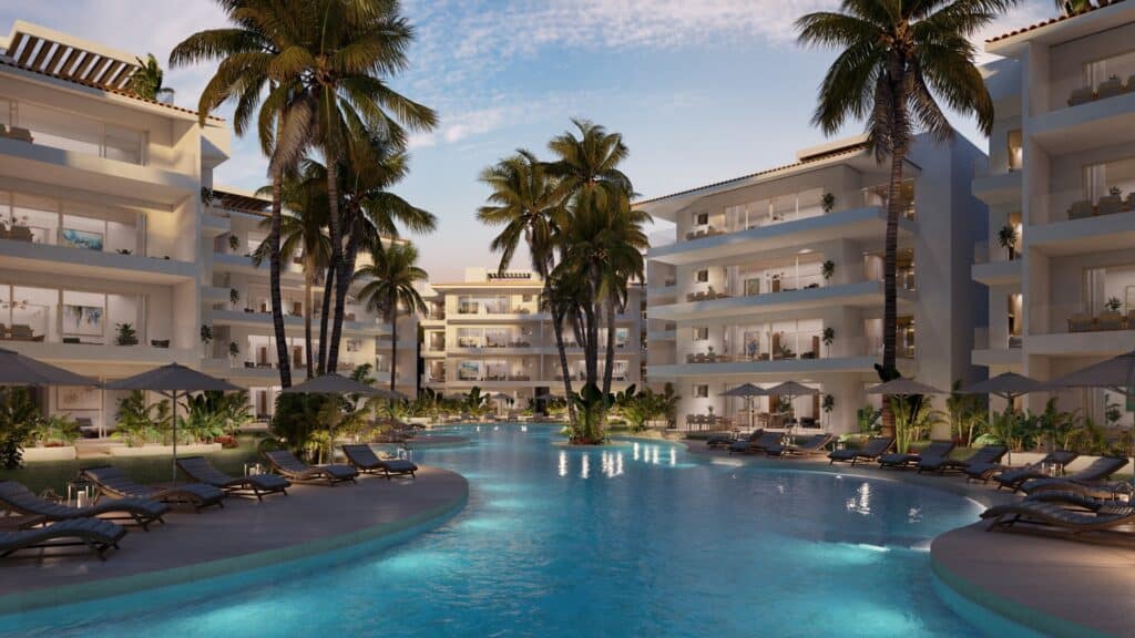 A pool area at Marina Aqua A in Puerto Aventuras, featuring a curving pool surrounded by lounge chairs, palm trees, and modern white residential buildings in a marina front setting.