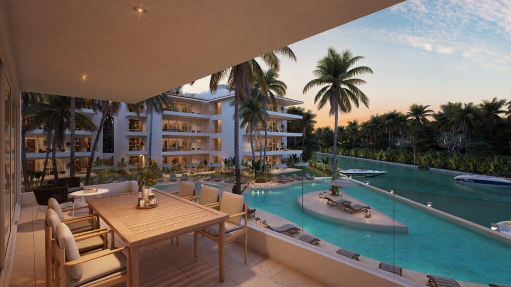 A spacious balcony overlooking a tropical pool area with palm trees at Marina Aqua A in Puerto Aventuras, showing yachts on the canal and a sunset sky.