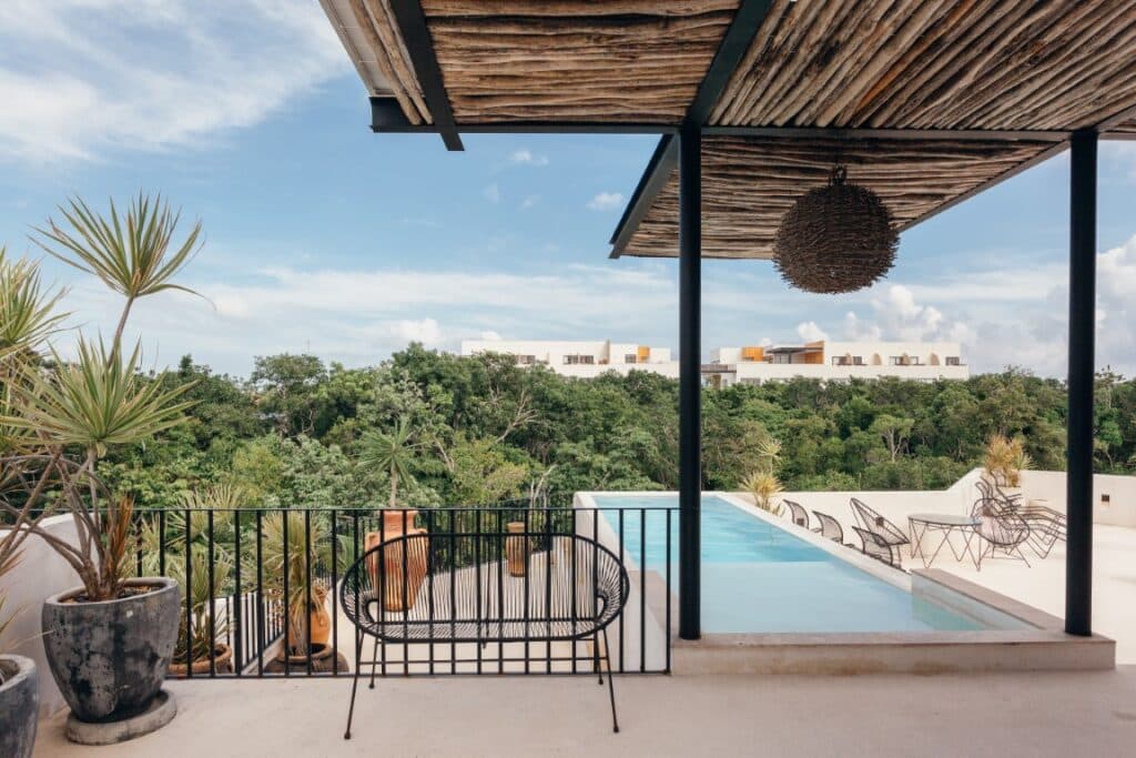 A view from Saastal in Aldea Zama, Tulum, showing a balcony with plants, a small pool, and outdoor seating, overlooking a lush green forest and modern buildings in the distance.