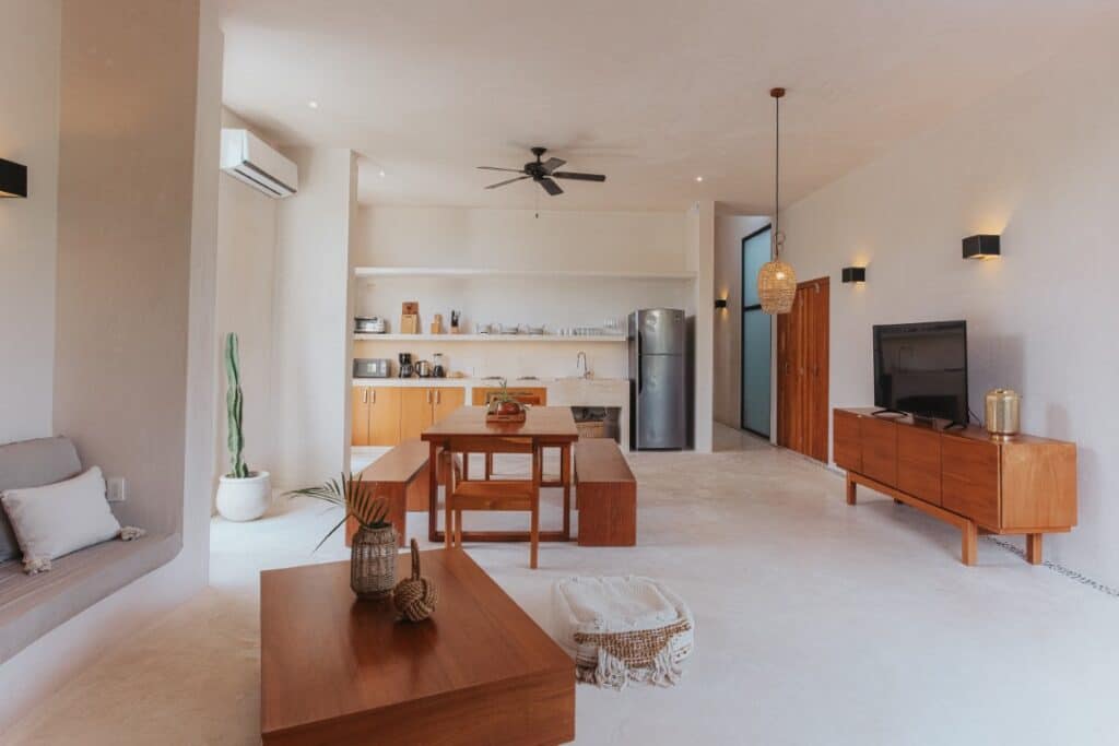 A spacious, modern interior at Saastal in Aldea Zama, Tulum, featuring a minimalist design with wooden furniture, a cozy sitting area, and an open kitchen layout.