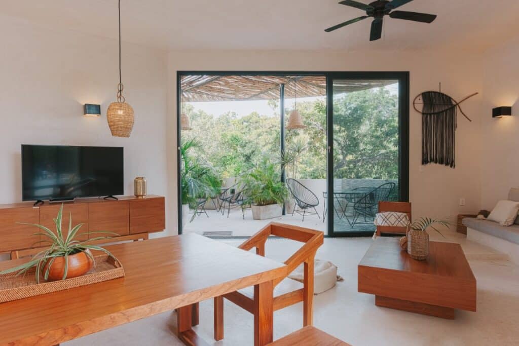 Interior view of a Saastal unit in Aldea Zama, Tulum, featuring a living area with wooden furniture, a sliding glass door leading to a balcony with outdoor seating and lush greenery.