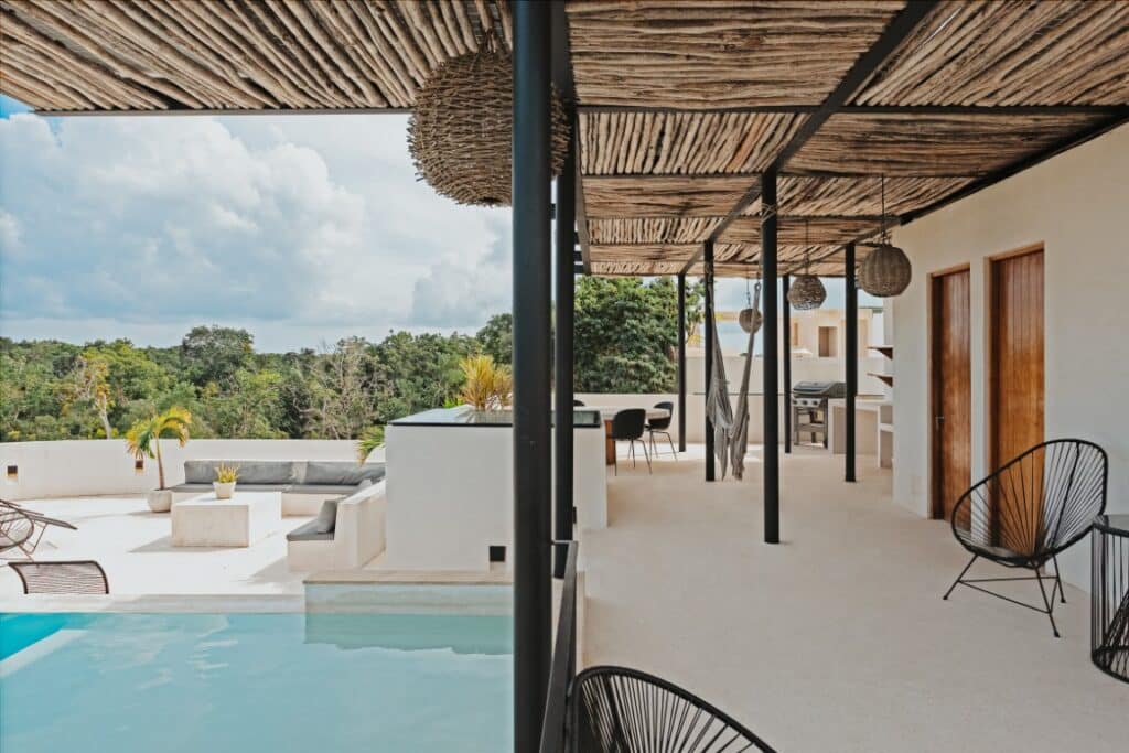 A spacious balcony at Saastal in Aldea Zama, Tulum, features a shaded area with seating, a hammock, and a view of lush green trees and the sky.