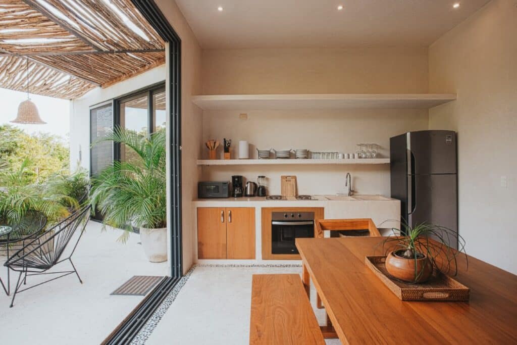 A modern kitchen in Saastal, Aldea Zama, Tulum, features a wooden dining table, open shelving with dishes, and sliding glass doors opening to an outdoor patio with greenery.