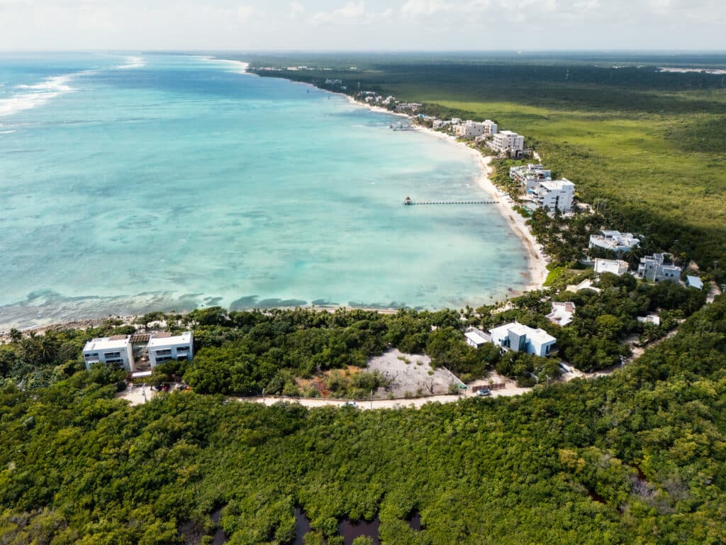 Aerial view of Tankah Bay Beachfront Land in Tulum, showing a stretch of white sandy beach with turquoise waters, modern white buildings, and lush green surroundings.