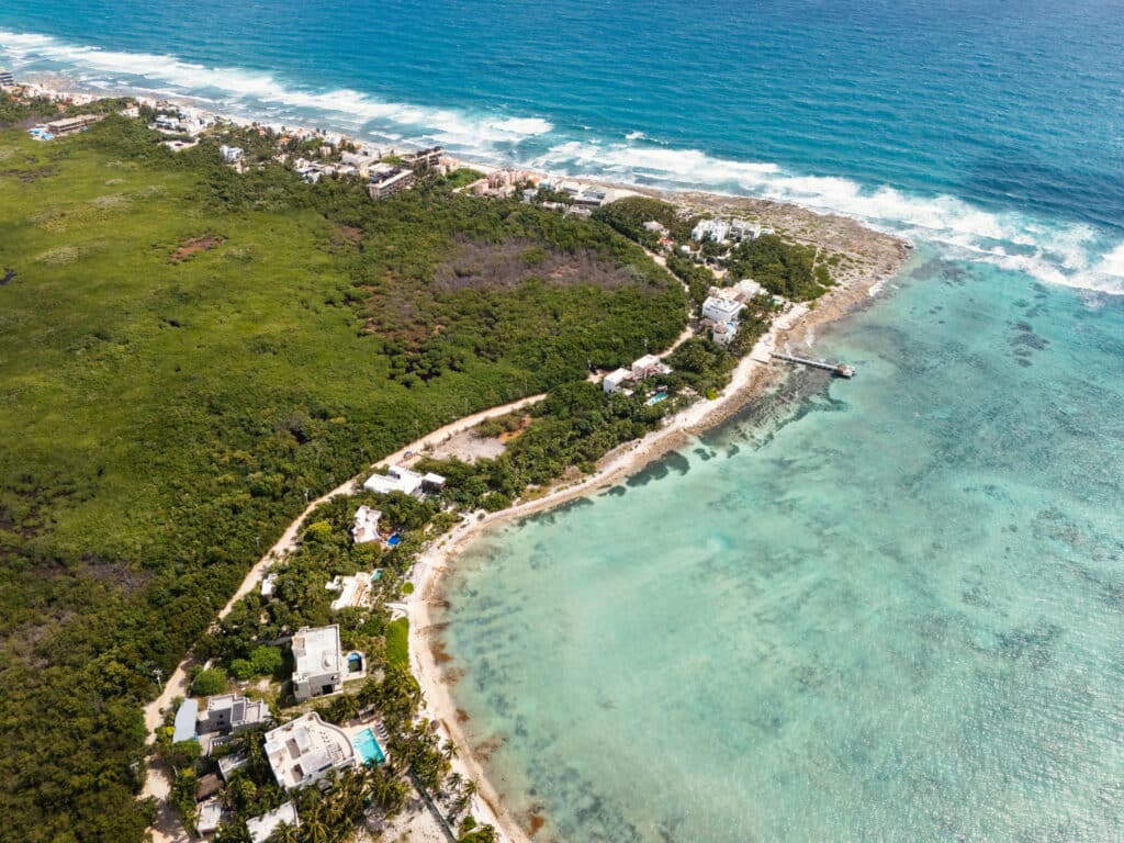 Aerial view of Tankah Bay Beachfront Land in Tulum showing lush green vegetation, beachfront homes, and a pier extending into the clear turquoise waters.